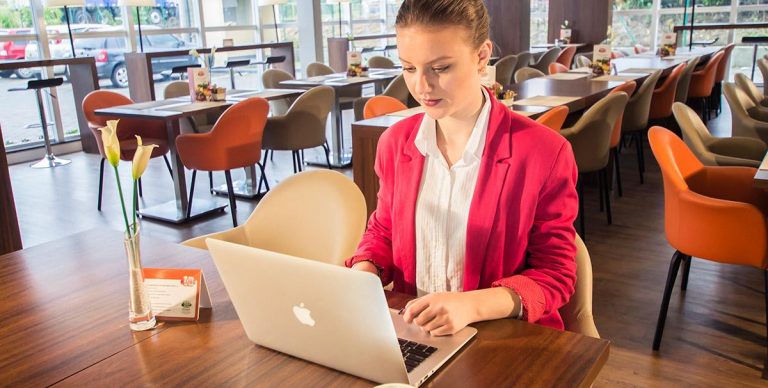 mulher de terno rosa sentada em uma mesa de convivío trabalhando em seu laptop